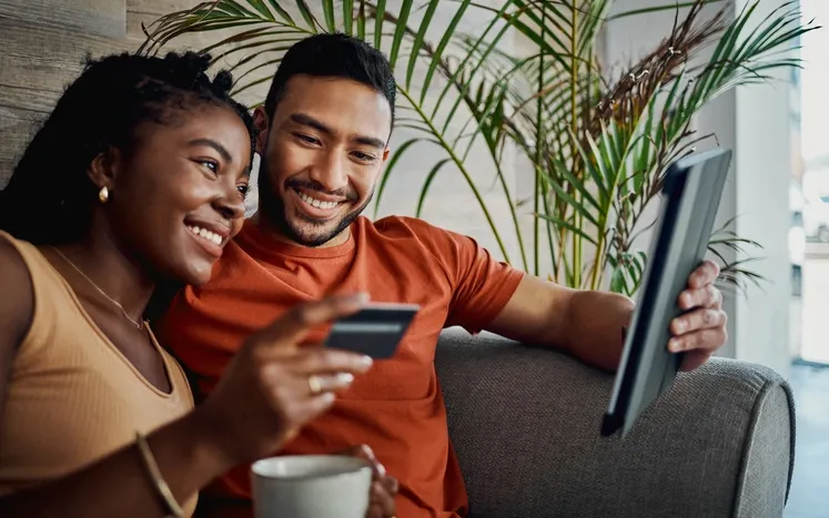 A couple is sitting on the couch while the girl holds the credit card, and the guy holds the laptop computer.
