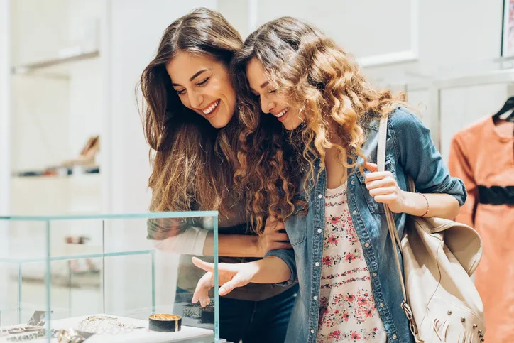 Two women are smiling and pointing to a display with jewelry inside.